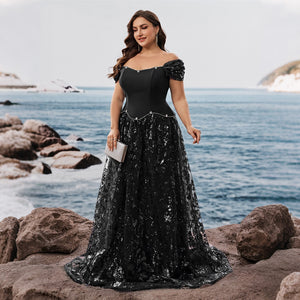 Curvy model showcasing a fitted black dress with a flowing skirt against a seaside backdrop. Perfect for cocktail attire and galas.